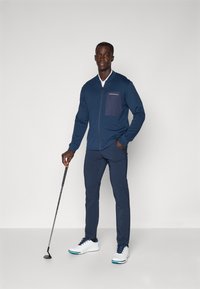 Marineblaue Zip-Jacke mit kontrastierendem Taschen, kombiniert mit marineblauen Hosen. Das Modell hält einen Golfschläger und trägt weiße Schuhe mit blauen Akzenten.