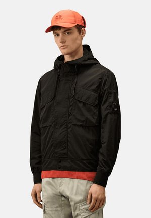 Giacca leggera nera con cappuccio, caratterizzata da due tasche frontali e dettagli a coste rosse all'orlo. Abbinata a un cappellino arancione.