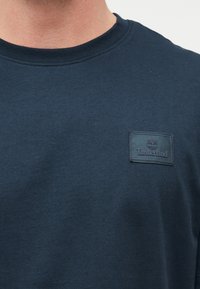 Κοντινό πλάνο ενός ατόμου που φοράει μπλε σκούρο t-shirt Timberland με ένα μικρό δερμάτινο ταμπελάκι λογότυπου στο στήθος.