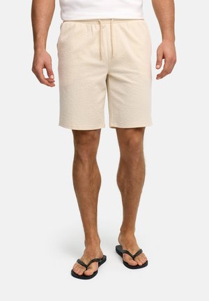 Mężczyzna noszący beżowe spodnie szorty z teksturą, z elastycznym pasem i sznurkiem, w czarnych klapkach, stojący na białym tle.