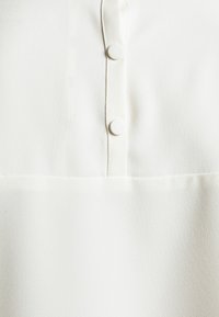 Vit blus med en slät, texturerad tyg, två runda knappar och en subtil fickdetalj nära fållkant.