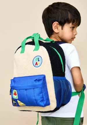 Tumejuustega laps, kes kannab valget särki ja rohelist püksipaarit, kannab musta ja kreemikat seljakotti roheliste rihmade ja sinise esitaskuga.