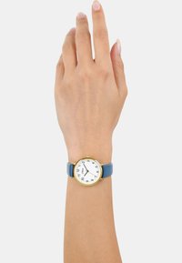 Goldfarbene Armbanduhr mit einem runden weißen Zifferblatt, arabischen Ziffern und einem hellblauen Lederarmband, die an einer Hand getragen wird.