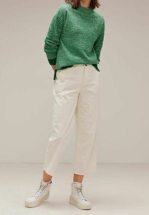 Personne portant un pull en tricot vert, un pantalon court taille haute écru et des baskets montantes blanches, debout devant un fond uni.