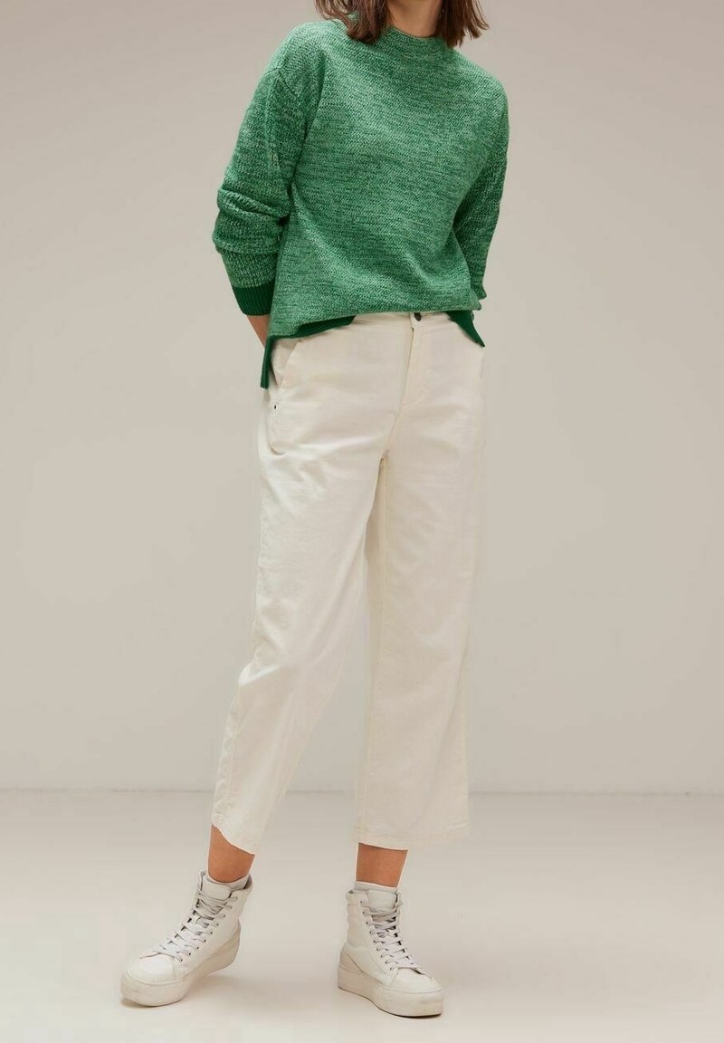 Personne portant un pull en tricot vert, un pantalon court taille haute écru et des baskets montantes blanches, debout devant un fond uni.