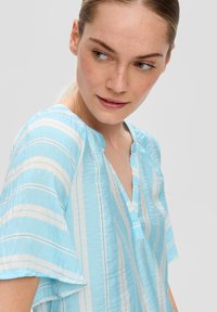 Chemise bleu clair en tissu texturé, dotée de rayures verticales blanches, d'un col en V et de manches courtes à coupe ample.