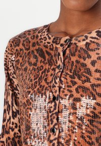 Leopardmønster cardigan i tan og brunt, med sorte knapper og perleaccenter, der fremhæver dyremønsteret. Blød tekstur.