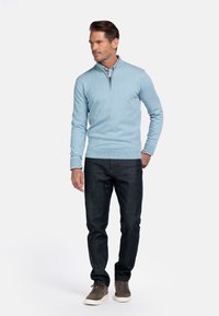 Lichtblauwe ritstrui van gebreide stof, gecombineerd met donkere jeans en grijze sneakers. Het witte shirt eronder heeft een bloemenpatroon.