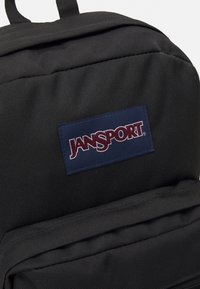 Črn najlon nahrbtnik z modrim pravokotnim flikom, na katerem je napis "JANSPORT" v činčani črti. Vključuje sprednji žep in oblazinjene naramnice.