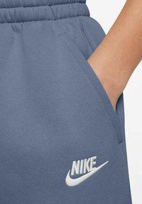 Harmaat Nike-housut, joissa on pehmeä rakenne, joustava vyötärö, sivutaskut ja valkoinen brodeerattu logo vasemmassa reidessä.