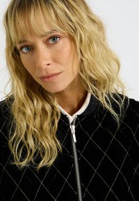 Veste bomber en velours noir avec couture en diamants argentés, dotée d'un col blanc et d'une fermeture éclair frontale. Le modèle a des cheveux blonds ondulés.