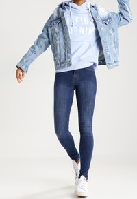 Chaqueta de mezclilla azul clara con detalles desgastados, superpuesta a una sudadera con capucha azul clara con texto. Jeans skinny azul oscuro y deportivas blancas.