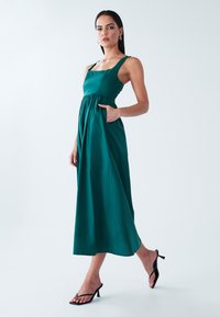 Teal pitkä mekko neliömäisellä kaula-aukolla, leveillä olkaimilla ja kerätyllä vyötäröllä, jossa on taskut. Yhdistetty mustiin hihnakenkiin vaalealla taustalla.