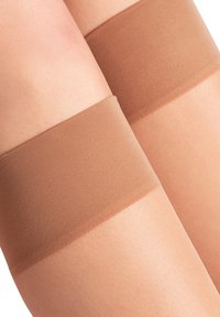 Dünne, hautfarbene Strümpfe in Oberschenkelhöhe mit einem breiten, festen Bund oben. Glatte Textur mit einem weichen Finish, entworfen, um sich eng an das Bein anzupassen.