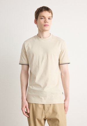 T-shirt beige à manches courtes en coton, avec des accents rayés noirs et blancs sur les manches et un col rond. Texture sans motif.