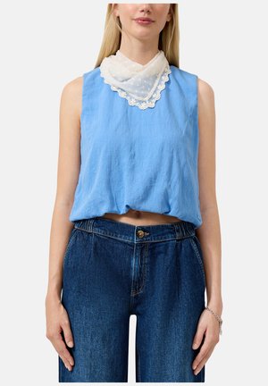 Femme portant un haut sans manches bleu clair, une écharpe blanche bordée de dentelle autour du cou, et un jean bleu foncé taille haute avec fermeture à boutons.