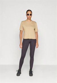 Top beige de manga corta con un logo blanco, combinado con leggings negros de corte ajustado y costuras visibles, usados con zapatillas negras.