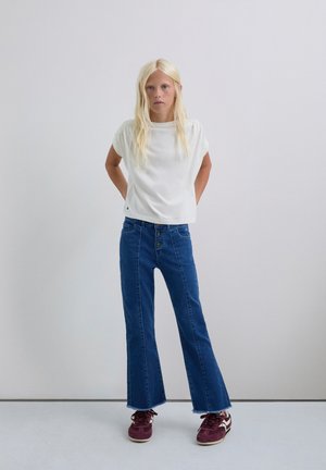 Top blanco de manga corta y jeans acampanados azules de talle alto con botones en el frente. Zapatillas rojas con acentos blancos completan el atuendo informal.