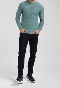 Maglietta a maniche lunghe a righe turchesi con scollo rotondo, abbinata a jeans scuri e scarpe sportive nere con accenti chiari.