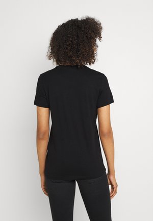Frau mit lockigem Haar, die ein schlichtes schwarzes T-Shirt mit kurzen Ärmeln und schwarze Hose trägt, steht mit dem Rücken zu einem einfarbigen hellen Hintergrund.