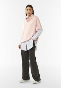 Hellrosa, übergroßer Strickpullover mit einem Rollkragen, über einem weißen Hemd mit Knöpfen getragen. Kombiniert mit dunkelgrauen, weiten Hosen und weißen Sneakers.