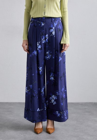 Pantalons larges floraux navy avec des motifs violets et bleus, présentant une texture lisse et une coupe ajustée, associés à des talons aiguilles marron.
