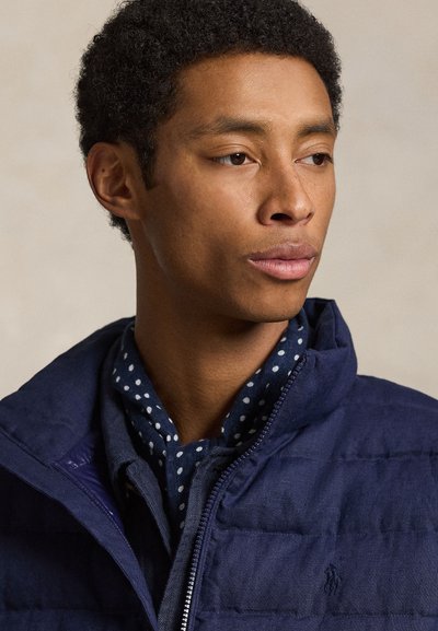 Młody mężczyzna o krótkich, kręconych włosach, ubrany w granatową, pikowaną kurtkę i niebieską koszulę w kropki, patrzący w prawo.