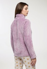 Pullover in pile viola, con texture e colletto alto, caratterizzato da un motivo a treccia. Indossato con pantaloni del pigiama chiari con stampa di orsetti.