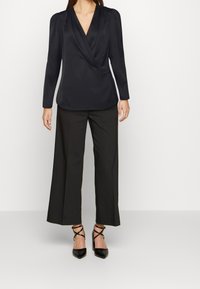 Langärmlige, schwarze Bluse mit tiefem V-Ausschnitt und gerafften Schultern, kombiniert mit weiten schwarzen Hosen und spitzen schwarzen Absätzen.