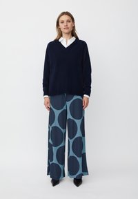 Granatowy sweter w serek zestawiony z szerokimi spodniami w kolorze teal, ozdobionymi dużymi ciemnoniebieskimi kółkami. Całość uzupełnia biała koszula z kołnierzykiem.