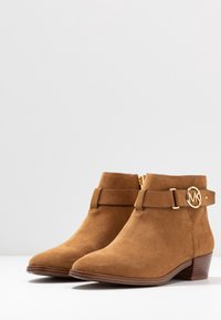 Brown suede ankle boots with a pointed toe, gold hardware accent, and a side zipper. Features a decorative strap with a circular logo.