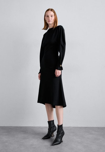 Black long-sleeve dress with fitted bodice and flared hem. Paired with pointed black ankle boots featuring textured detailing on heels.