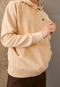 Hoodie beige chiaro con texture a macchie, tasca a marsupio, stringhe regolabili e logo ricamato sul petto.
