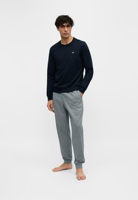 Jeune homme debout pieds nus portant une chemise noire à manches longues et un pantalon de jogging gris à motifs sur un fond blanc uni.