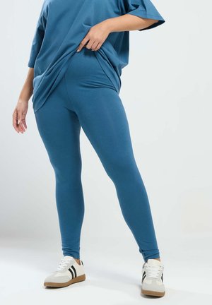 Osoba w niebieskich legginsach i za dużym niebieskim topie, lekko podciągająca koszulkę, w białych trampkach z czarnymi paskami.