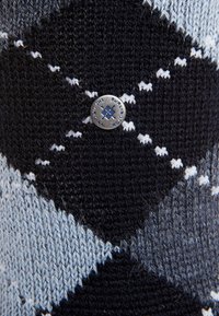 Primer plano de una tela de punto con un patrón de argyle en negro, azul claro y blanco, que presenta un pequeño botón de metal redondo etiquetado "Burlington".