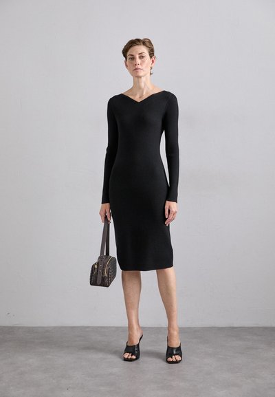 Black ribbed long-sleeve dress with a V-neck, fitted silhouette, knee-length hem, and smooth texture. Model carries a small patterned handbag.