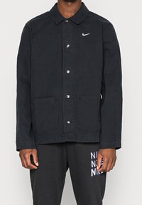 Svart jacka med knappar och krage, två framfickor och en liten vit Nike-logotyp på bröstet. Tillverkad av ett slitstarkt tyg.