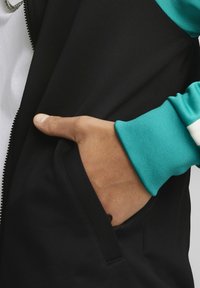 Svart zip-up jacka med turkosa ärmar. Har en ficka fram med en hand som vilar inuti, vilket visar en slät textur.