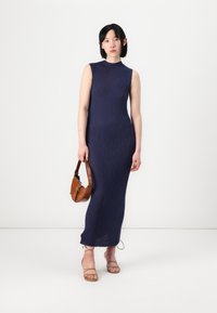 Rochie fără mâneci, de culoare bleumarin, cu un finisaj texturat, lungime până la gleznă, fante laterale și tiv cu ușoare destrăbălări. Asortată cu sandale cu brățări și o geantă maro.
