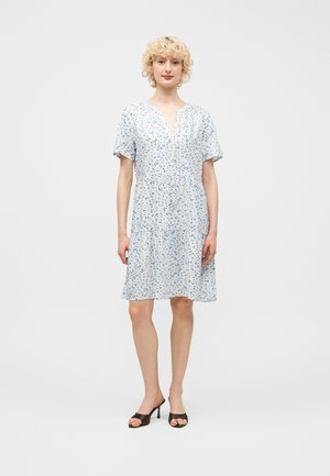 Žena s kučeravými blond vlasmi, oblečená v bielych kvetovaných šatách s krátkym rukávom a čiernych sandáloch s odkrytými prstami, stojaca pred jednoliate pozadie.