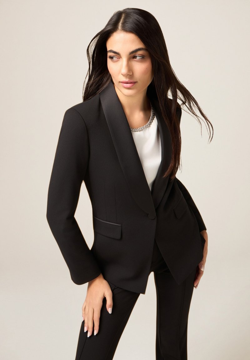 Czarna, dopasowana marynarka z satynowymi klapami, o fasonie dopasowanym i kieszeniach po bokach. Noszona na białym topie z metalicznym ozdobieniem przy dekolcie.