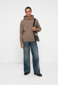 Sudadera marrón con logo en relieve, vaqueros azules de corte holgado y zapatos negros. Accesorio: pequeña bolsa negra de hombro con correa.