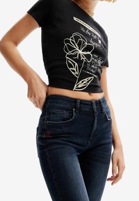 Top negro de canalé de manga corta con gráfico floral blanco y texto; combinado con jeans de tiro alto azul oscuro con bordado de acento rojo.