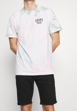Osoba ubrana w białą i pastelową koszulkę tie-dye marki Levi's oraz czarne szorty, stojąca z rękami za plecami na białym tle.