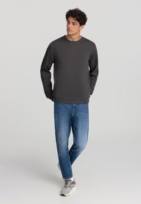Felpa grigia con scollo a giro, abbinata a jeans azzurri chiari. Il modello indossa sneakers grigie e bianche. Design semplice, senza motivi visibili.