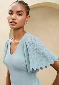 Top tricotat de culoare bleu deschis, cu decolteu în V, corp cu nervuri și mâneci bufante cu margini ondulate, decorate cu un model texturat. Design minimalist și țesătură moale.