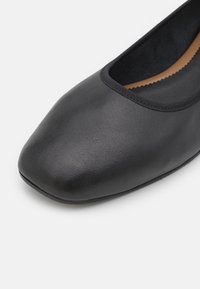 Gros plan sur une chaussure plate noire avec un bout carré arrondi et une surface subtilement texturée sur fond blanc.