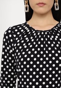Black blouse with white polka dots, featuring a gathered neckline and long sleeves. Smooth fabric with a regular fit. Statement earring visible.