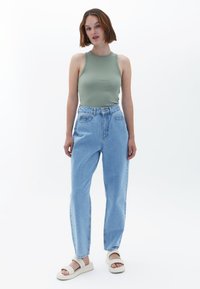 Top verde, jeans in denim azzurro a vita alta, vestibilità rilassata, tessuto morbido, abbinati a sandali bianchi a infradito su uno sfondo neutro.